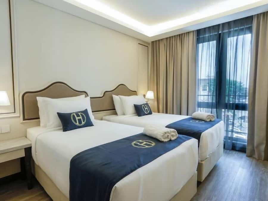 A hotel room with two beds, each with a headboard, bedside tables with lamps, and a view of the outdoors through large windows.