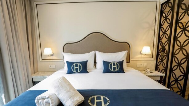 A hotel room with a bed, two pillows with a logo, a bedside table, and a wall with a decorative panel. - THE HOST HOTEL (Ipoh)