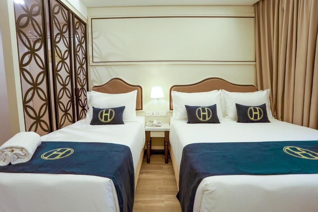 A hotel room with two beds, each with matching headboards and pillows, a nightstand with a lamp, and a decorative panel on the left.