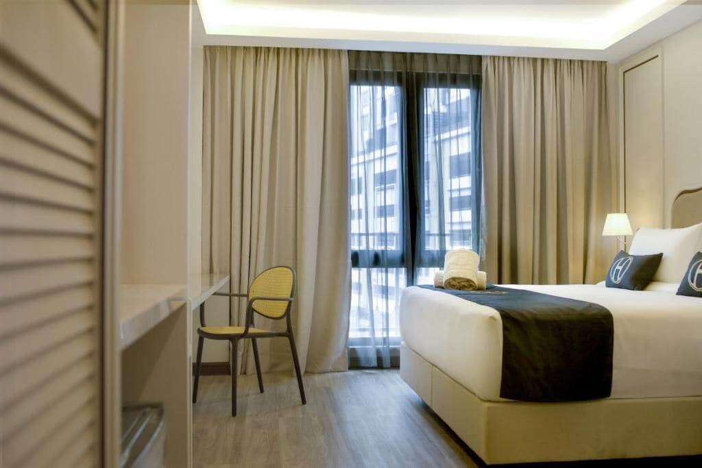 Habitación de hotel con cama, una silla, mesita de noche, lámpara y ventana con cortinas.