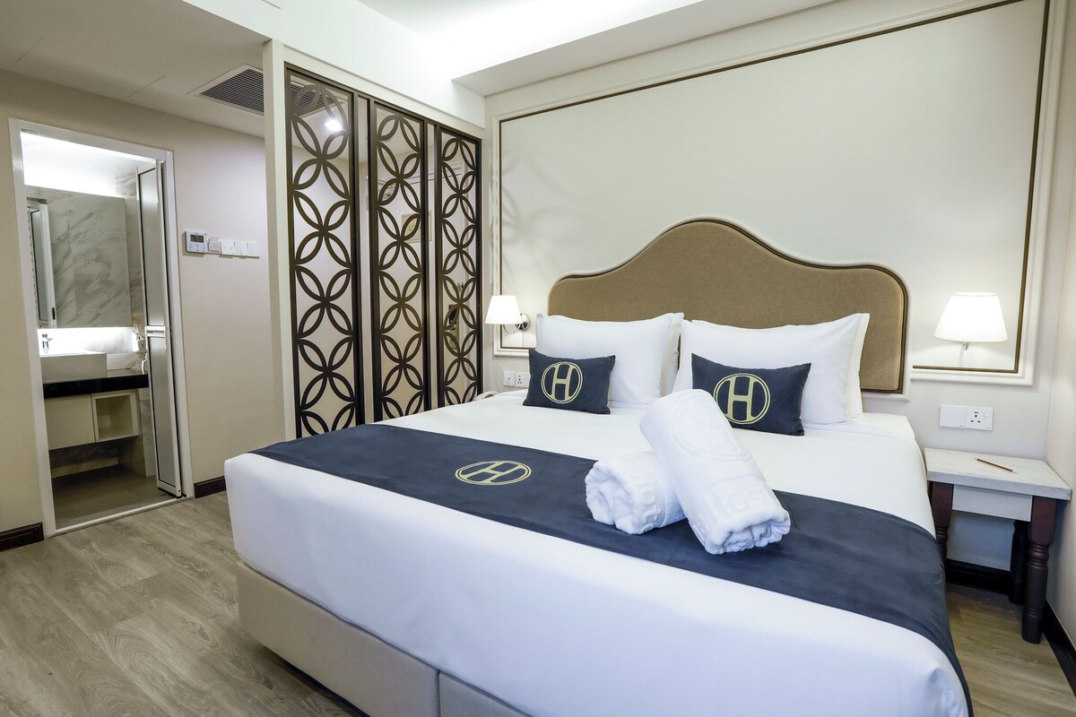 Habitación de hotel con cama, mesita de noche, lámpara, baño con espejo y un biombo decorativo.
