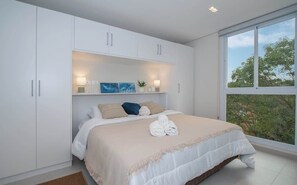 Un dormitorio con una cama grande, armarios blancos, una ventana con vistas y un estante con adornos.