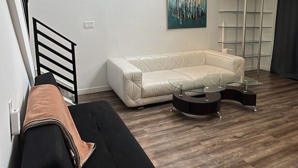 A modern living room with a black sofa, a white couch, a glass coffee table, a painting on the wall, and a wooden floor.