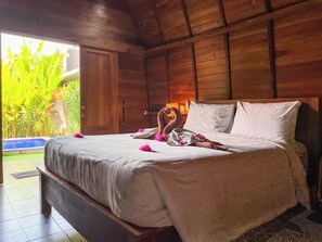 A wooden room with a bed, a view of greenery outside, and a door. - Jineng Villa Sanur 1 (Denpasar selatan)
