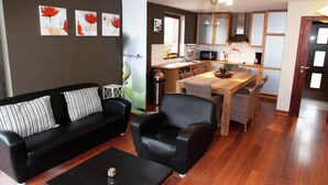 Uma sala de estar moderna com um sofá de couro preto, uma mesa de jantar com cadeiras e uma área de cozinha ao fundo.