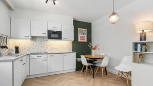 Moderni keittiö, jossa on valkoiset kaapit, kalanruotokuviollinen lattia, ruokapöytä tuoleineen ja kirjahylly.