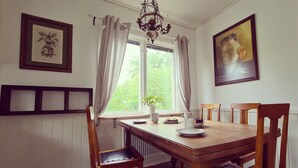 Uma sala de jantar com mesa de madeira, cadeiras, um candelabro, janela com cortinas e quadros nas paredes.