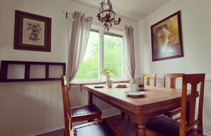 A dining room with a wooden table, chairs, a chandelier, a window with curtains, and framed artwork on the walls. - Vacation home in the idyllic village of Bolmen, 500 meters from Lake Bolmen (Ljungby)