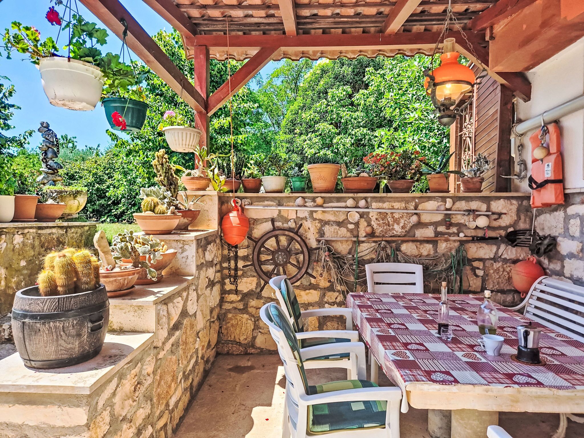 An outdoor patio with a stone wall, a table with a patterned tablecloth, chairs, potted plants, and hanging lights.
