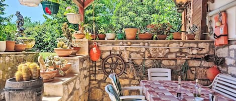 An outdoor patio with a stone wall, a table with a patterned tablecloth, chairs, potted plants, and hanging lights.