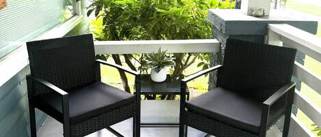 A covered outdoor seating area with two chairs and a table, surrounded by greenery and a view of a house.