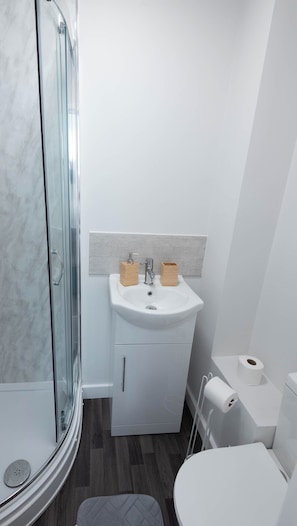 A bathroom with a glass shower enclosure, a white sink with wooden soap holders, a toilet, and a small storage cabinet. - The Bay Apart-Hotel by Guestz (Morecambe)