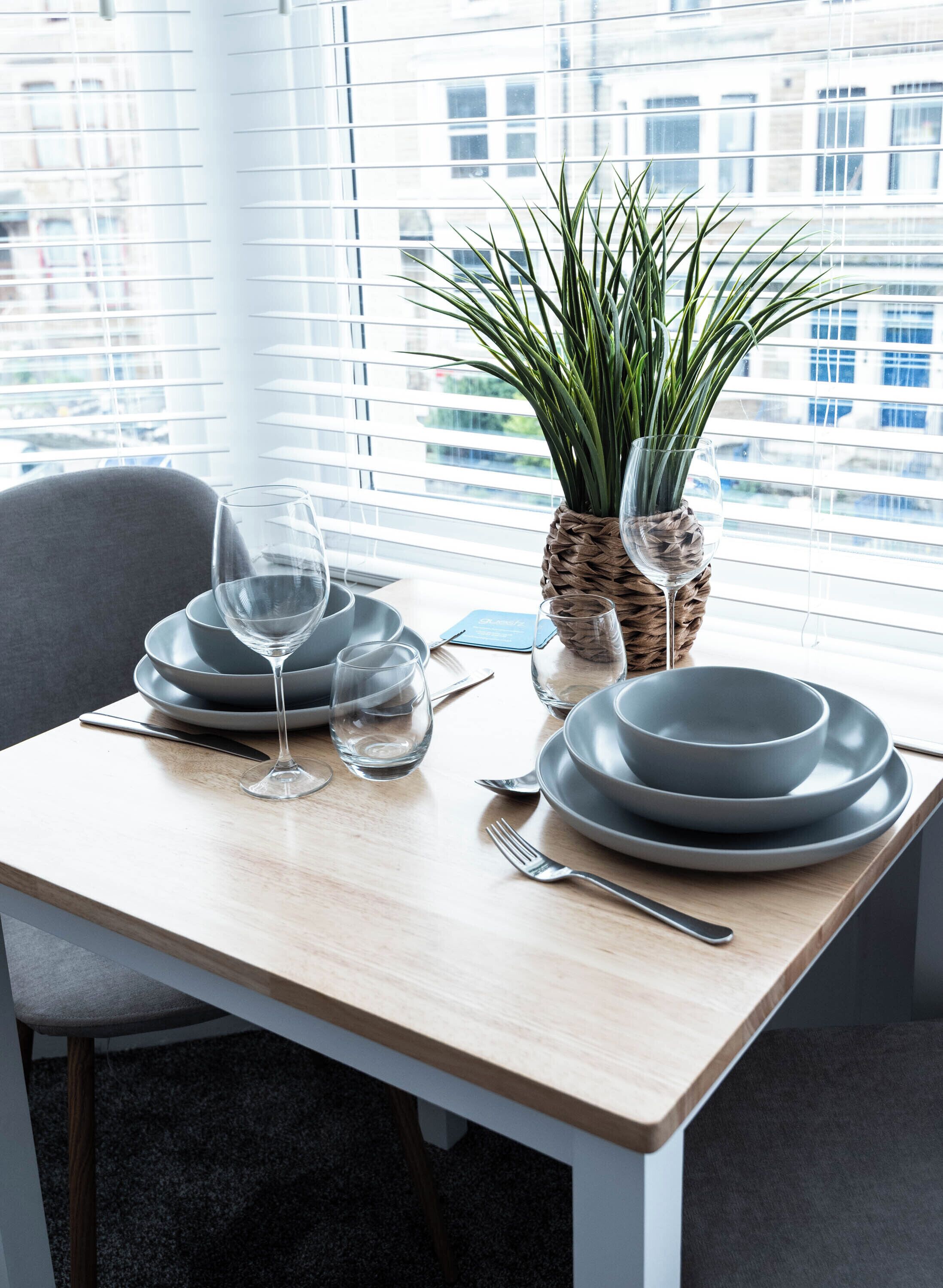 Una mesa de comedor dispuesta con platos, vasos y cubiertos, una planta en maceta y un teléfono inteligente sobre la mesa.