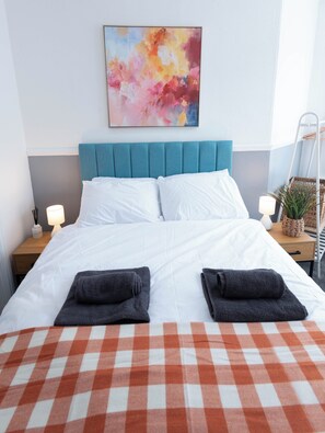 A neatly made bed with white linens and two folded towels, a teal headboard, and a colorful abstract painting above. - The Bay Apart-Hotel by Guestz (Morecambe)