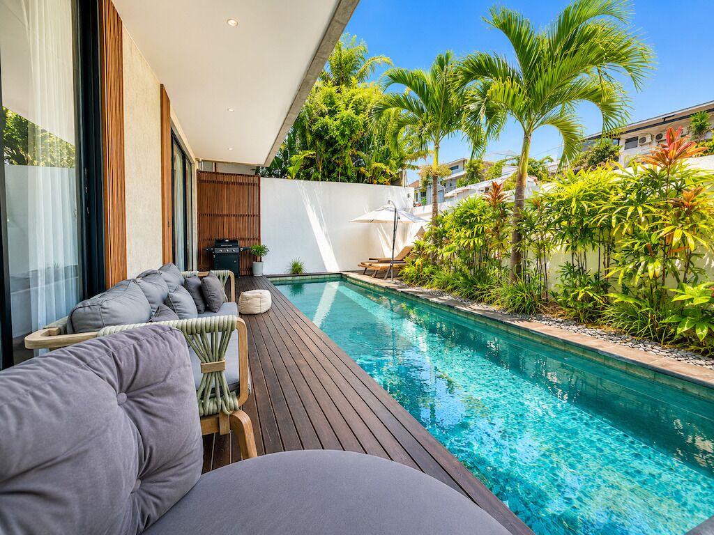 A modern poolside area with a wooden deck, a seating area with cushioned chairs, and a pool surrounded by tropical plants.