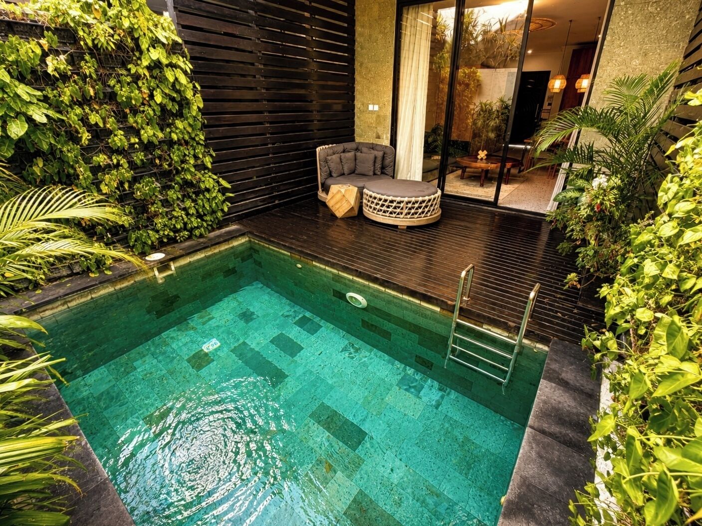 A swimming pool with a wooden deck, surrounded by lush greenery and a modern interior.
