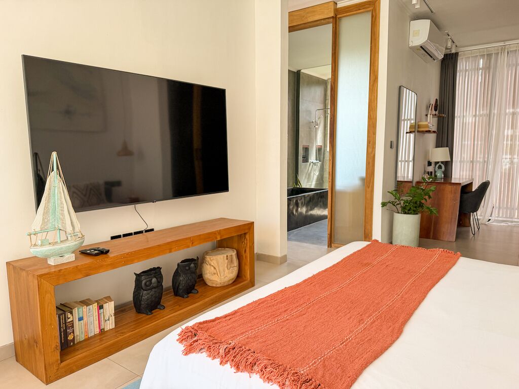 A hotel room with a large flat-screen TV, a wooden console table with books and decorative items, a large bed with an orange throw, and a view of the bathroom and balcony.