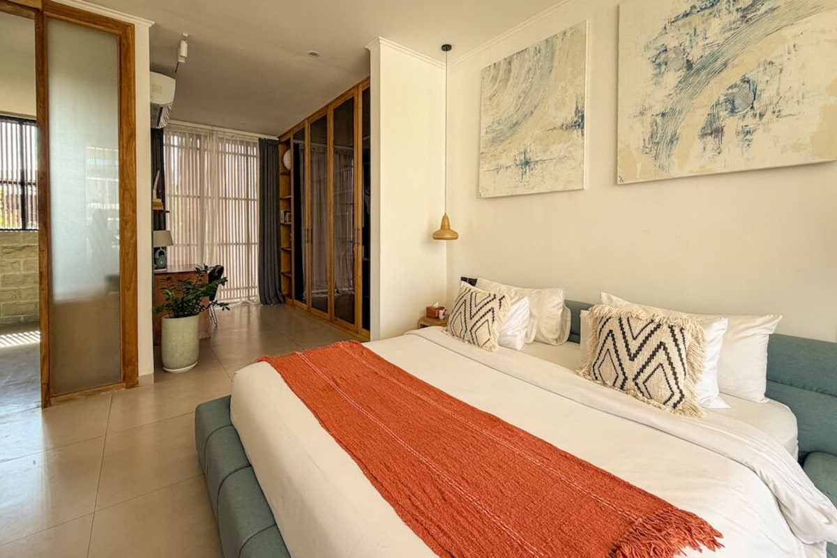 A bedroom with a large bed, a bedside table, a lamp, and abstract wall art.