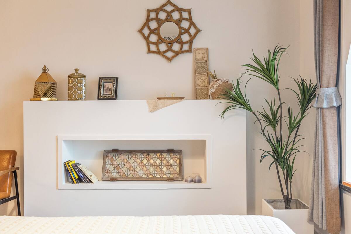 Un dormitorio con una cama blanca, una estantería con adornos, un espejo de pared y una planta en maceta.