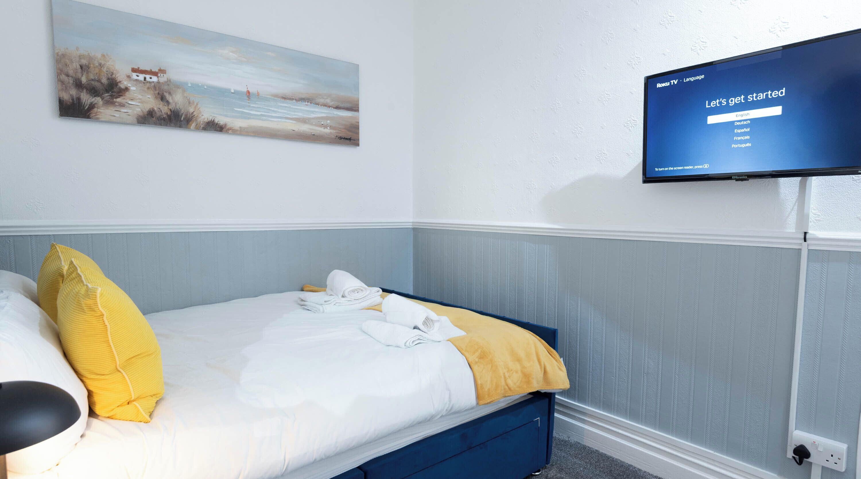 A hotel room with a bed, pillows, a painting, and a TV displaying a language selection screen.