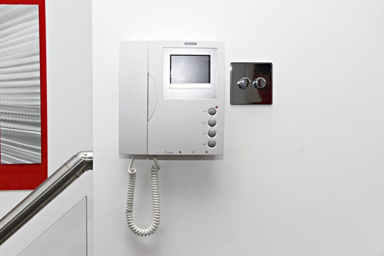 Een aan de muur bevestigde telefoon met scherm en knoppen, een opgerolde kabel en een bedieningspaneel aan de muur.