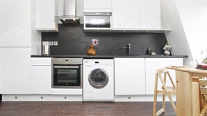 Uma cozinha moderna com armários brancos, forno embutido, máquina de lavar roupa e área de refeições com mesa e cadeiras de madeira.