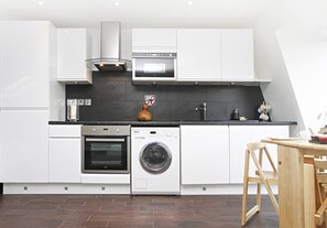 Una cocina moderna con armarios blancos, horno integrado, lavadora y comedor con mesa y sillas de madera.
