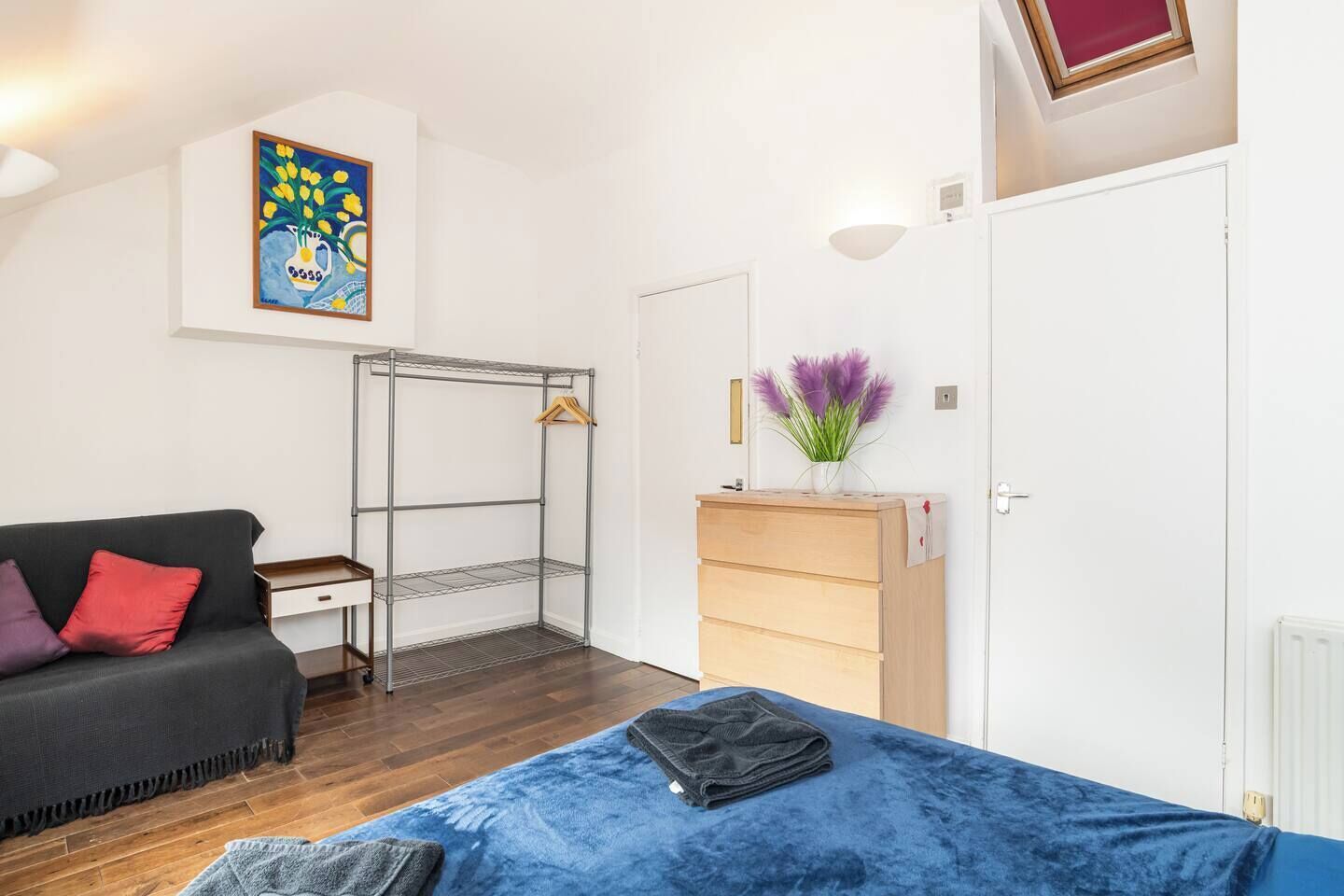 Une pièce comprenant un lit, un canapé, une armoire en métal, une commode, un radiateur, un tableau et une fenêtre.