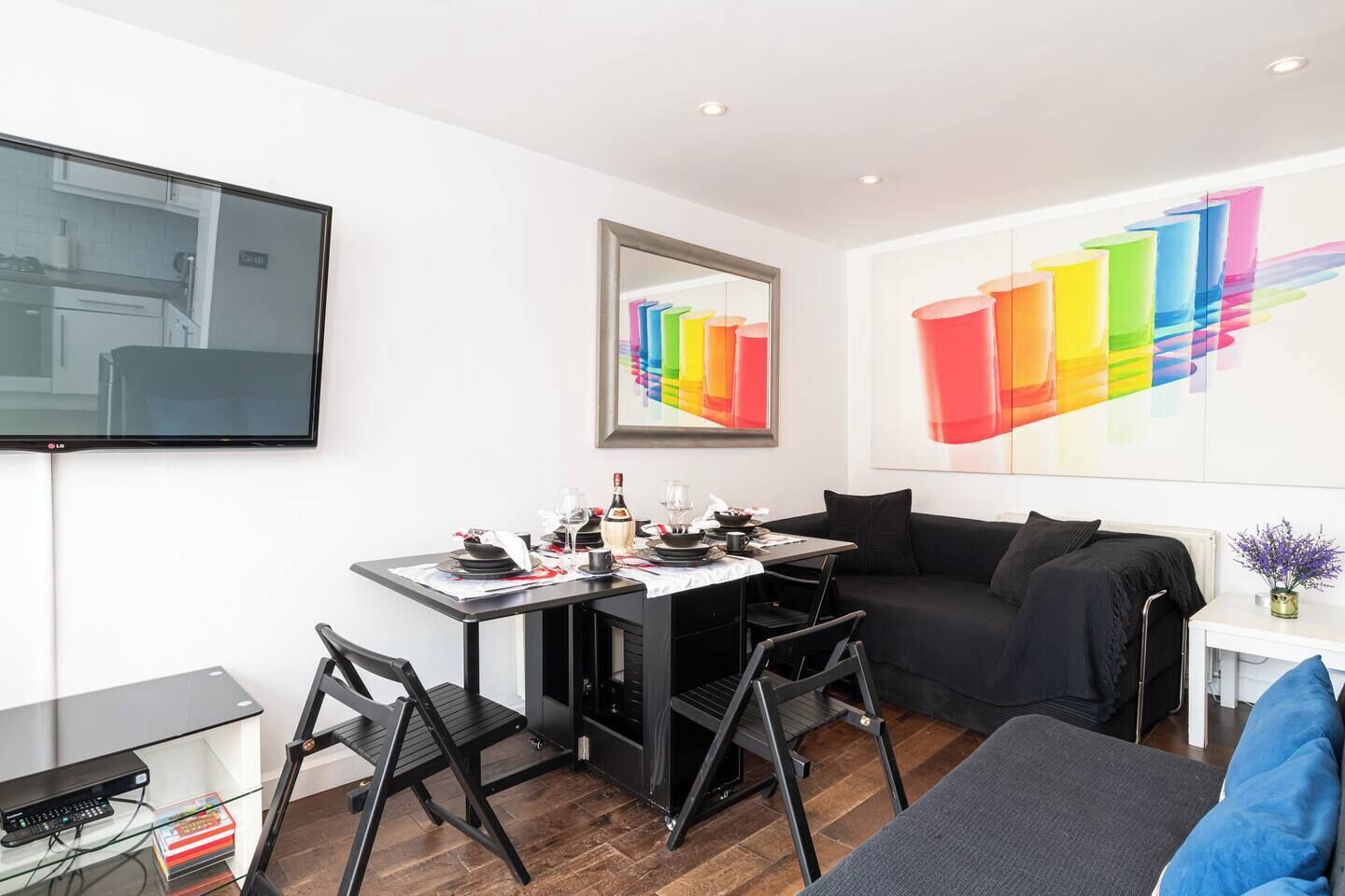 Ein modernes Wohnzimmer mit einer schwarzen Couch, einem Esstisch für zwei Personen und einem an der Wand befestigten Fernseher.