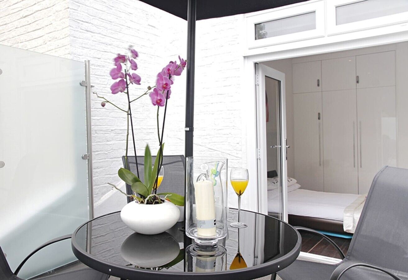 A glass table with a white vase, purple orchids, a candle, and a glass of orange juice.