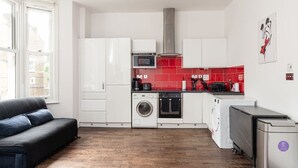 Eine kleine Küche mit weißen Schränken, einer roten Spritzschutzwand, einer Waschmaschine und einem kompakten Kühlschrank.
