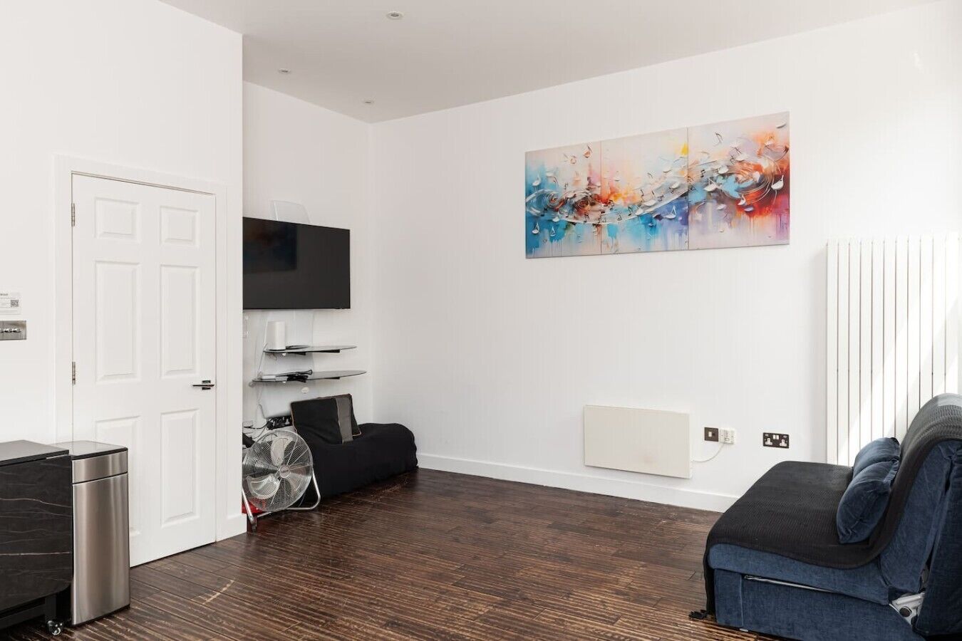 A modern living room with a flat-screen TV, a black sofa, a standing fan, and a large abstract painting on the wall.