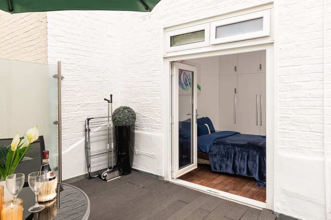 Ein Zimmer mit einem Bett, einer Tür zu einem anderen Raum, einem kleinen Tisch mit einer Vase und Gläsern sowie einem Fenster mit Jalousien.