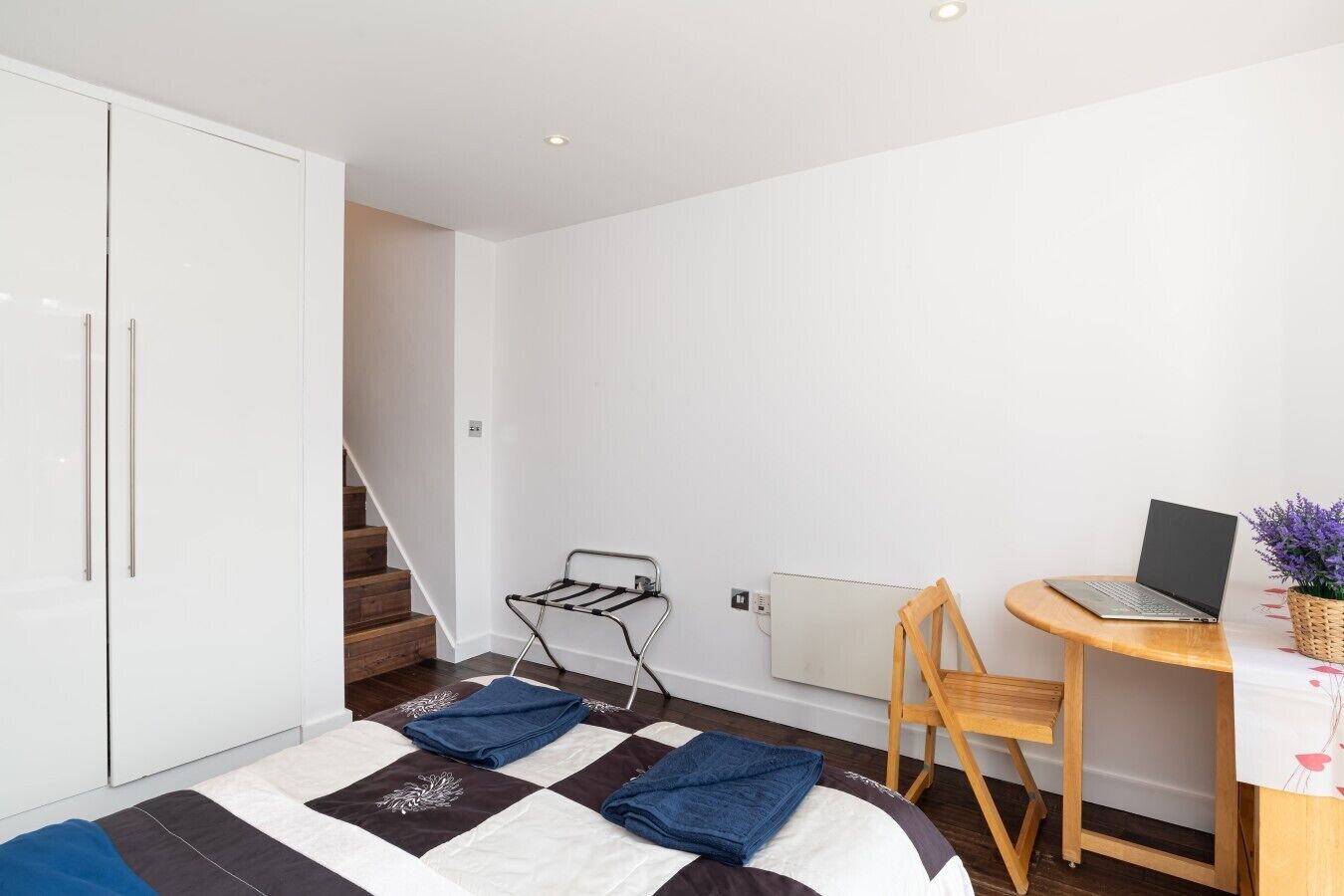 A bedroom with a bed, a desk with a laptop, a radiator, and a staircase.