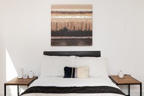 Une chambre à coucher avec un lit, deux tables de chevet, une œuvre d’art accrochée au mur et une fenêtre qui projette une ombre.