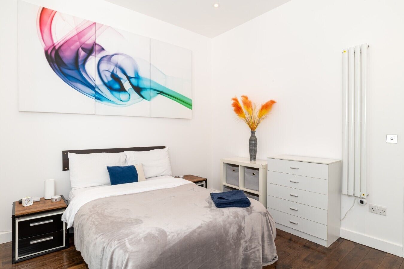 Ein modernes Schlafzimmer mit einem Bett, Nachttischen, einer Kommode, einem Heizkörper und einem großen abstrakten Gemälde.