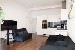 A modern living room with a flat-screen TV, a sofa, and a couch. There is a kitchen area with white cabinets and a microwave. - 2BR Family Garden Flat Sleeps 8 (Greater London)