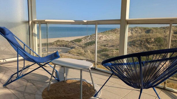 Ein Balkon mit Blick auf Strand und Dünen, mit zwei Stühlen und einem kleinen Tisch.
