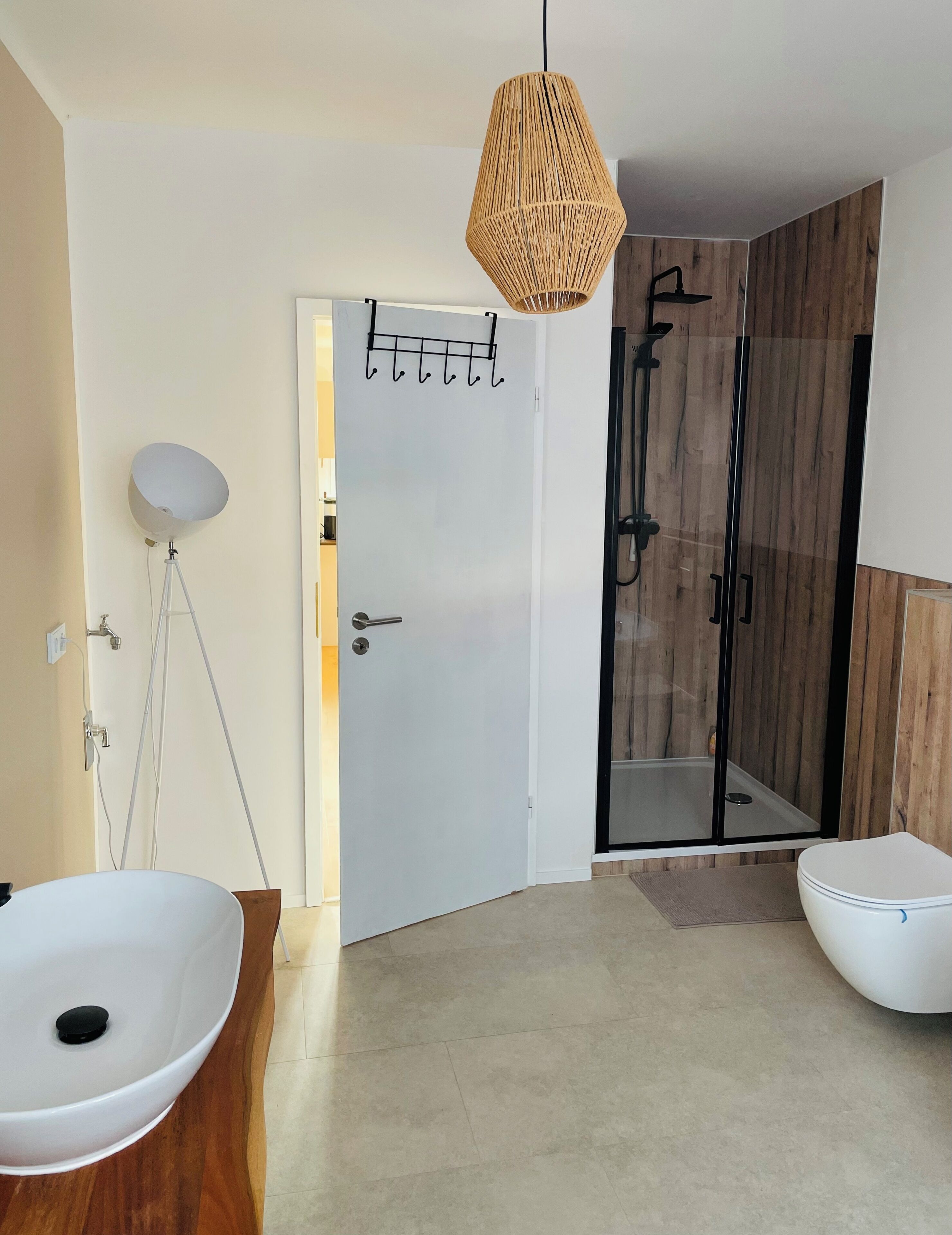 Un baño moderno con un lavamanos grande blanco, un espejo redondo, una puerta blanca con toallero y una ducha con puerta de cristal. Se observa un colgador de luces.