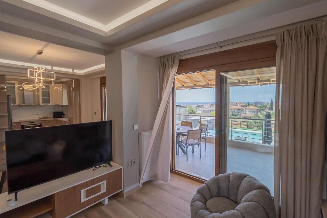 A modern living room with a flat-screen TV, a dining area with a table and chairs, and a balcony with a view of the sea.