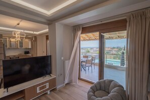 A modern living room with a flat-screen TV, a dining area with a table and chairs, and a balcony with a view of the sea.