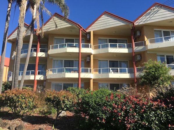 A multi-story building with balconies, brick exterior, and surrounding vegetation.