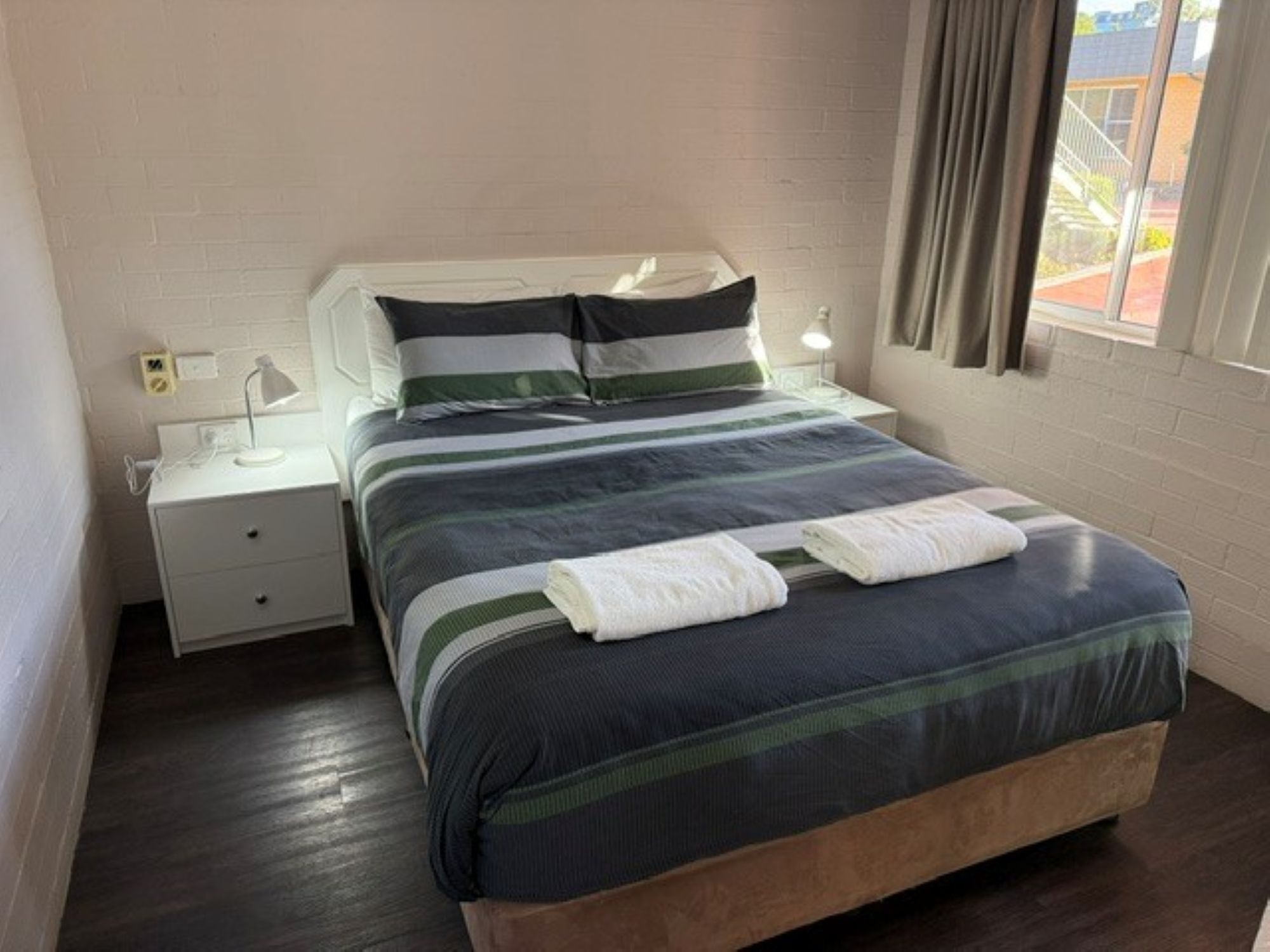 Una cama bien hecha con una colcha a rayas, una lámpara de noche y una ventana con cortinas.