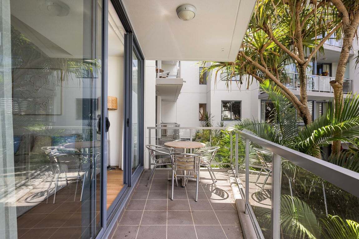 Un balcón con puerta de vidrio, una mesa redonda con sillas y vista a un edificio con balcones y plantas.
