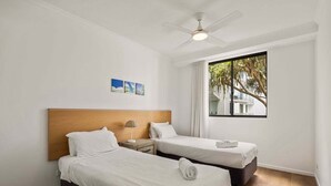 Una habitación de hotel con dos camas, un ventilador de techo, una ventana con vistas a palmeras y una pequeña mesa con una lámpara.
