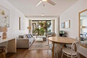 Ein modernes Wohnzimmer mit Sofa, Couchtisch, Esstisch und Stühlen. Es gibt einen Deckenventilator, einen Fernseher und Kunstwerke an den Wänden.