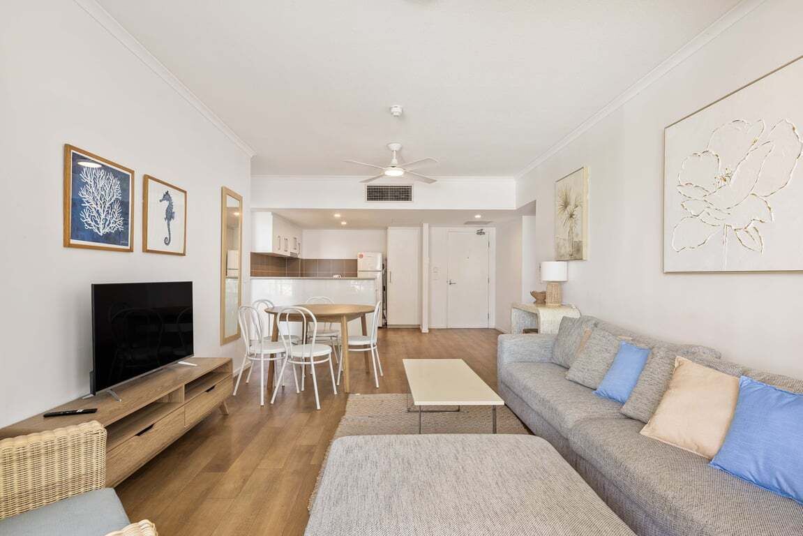 A modern living room with a flat-screen TV, a sofa, a dining table with chairs, and a kitchen area in the background.