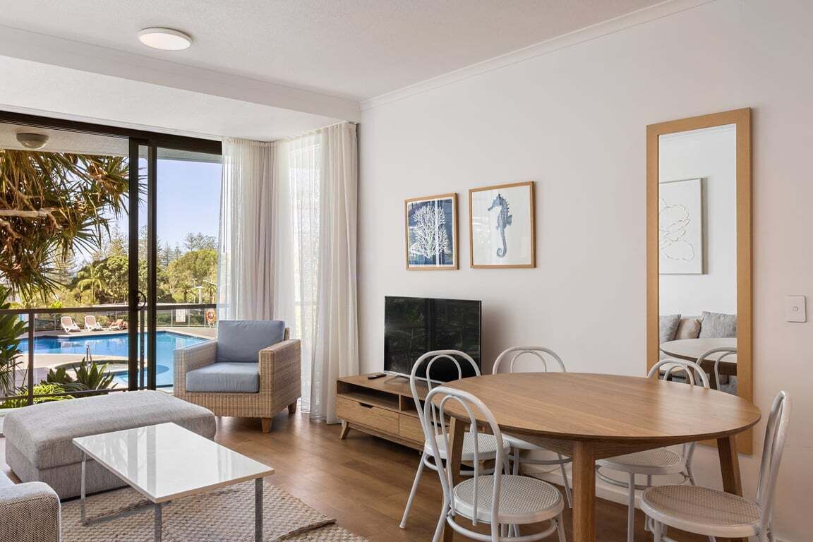 Una sala de estar moderna con una mesa de comedor de madera, sillas blancas, un sofá y un televisor. Hay cuadros enmarcados en la pared y un ventanal con vistas a una piscina.