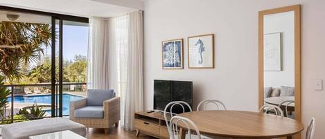 Una sala de estar moderna con una mesa de comedor de madera, sillas blancas, un sofá y un televisor. Hay cuadros enmarcados en la pared y un ventanal con vistas a una piscina.