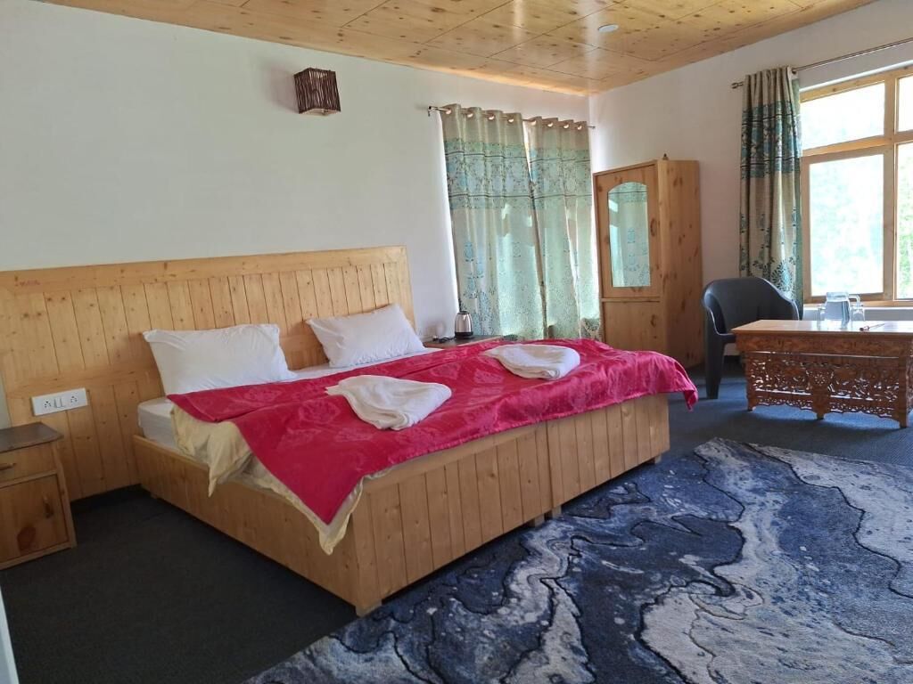 A wooden bed with white pillows and a red blanket, a wooden nightstand, a desk with a chair, and a window with curtains.